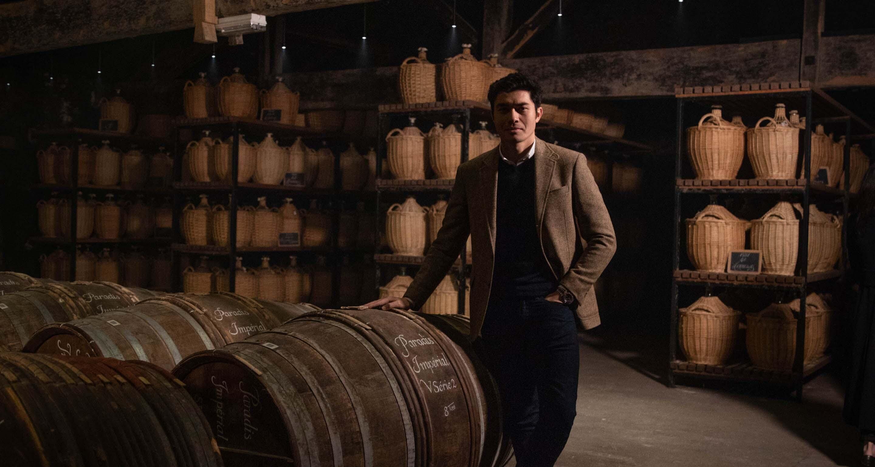 Henry Golding stands among the stacked casks of Hennessy cognac in the aging cellar.