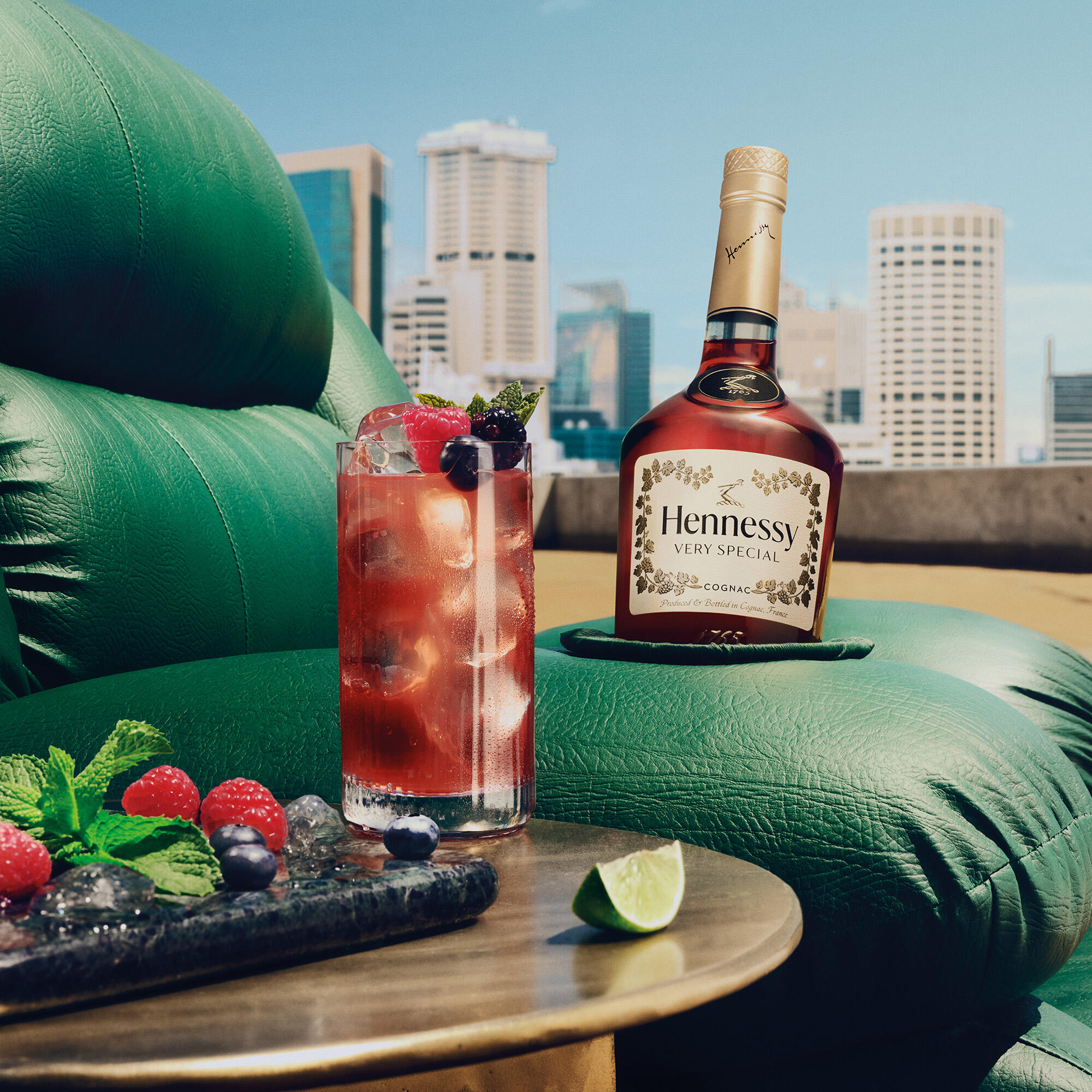 Hennessy V.S. bottle resting on a plush green couch with a refreshing Berry Mojito and a platter of berries on a side table.